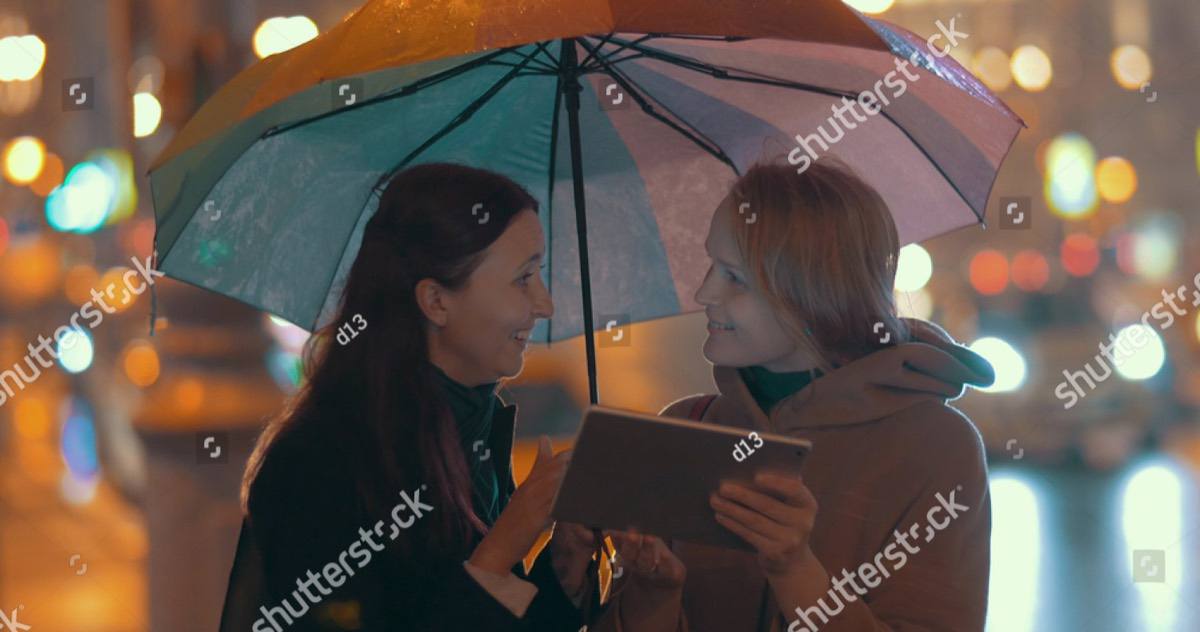 Two women sharing one umbrella at night, talking, lit by warm city lights